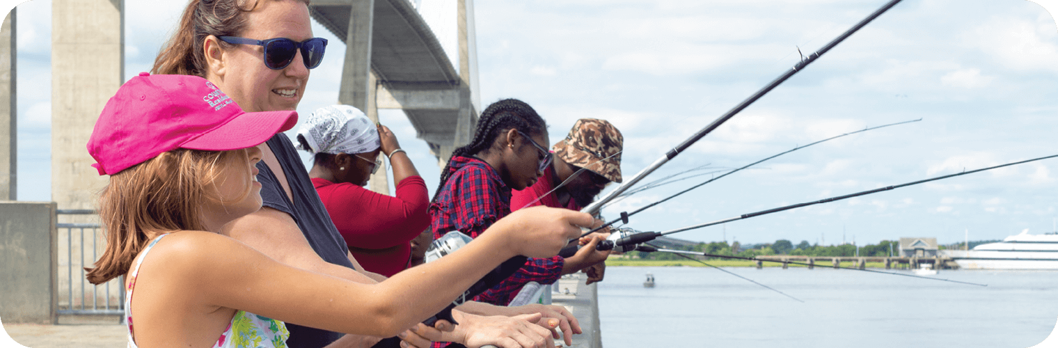 A diverse group of people, including children and adults, stand along a waterfront pier under a large bridge, fishing with rods over the railing. A young girl in the foreground wearing a bright pink hat and colorful tank top smiles as she holds a fishing rod with assistance from an adult woman in sunglasses. The background features calm water, a boat in the distance, and a partly cloudy sky.