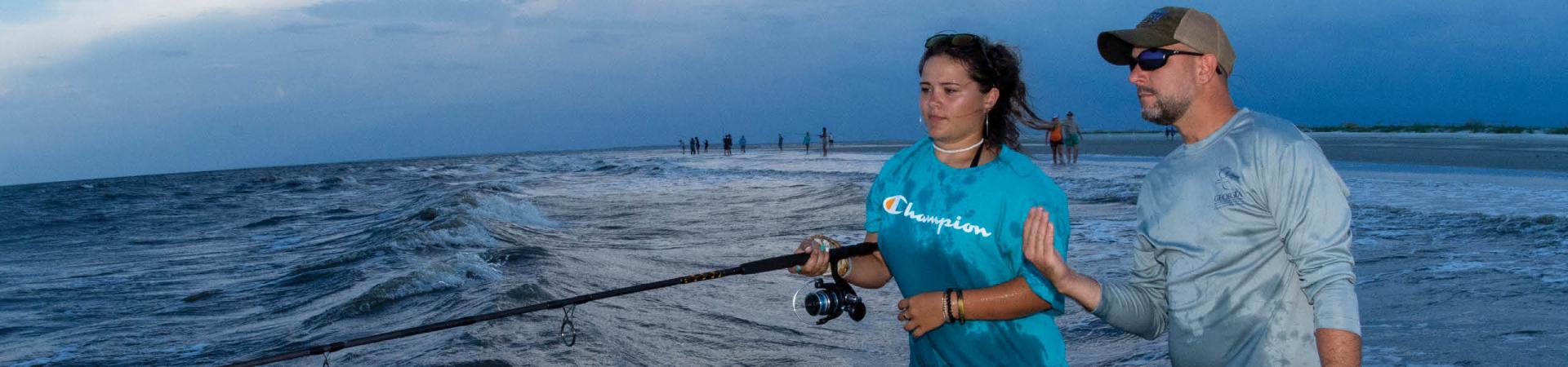 A young woman wearing a blue t-shirt and bracelets holds a fishing rod while standing in the surf at the edge of the ocean. Beside her, a man in a light gray long-sleeve shirt, sunglasses, and a cap provides guidance, gesturing with his hand. The waves roll onto the shore under a darkening sky, suggesting early evening or dusk. In the background, several other people are fishing and walking along the shoreline.