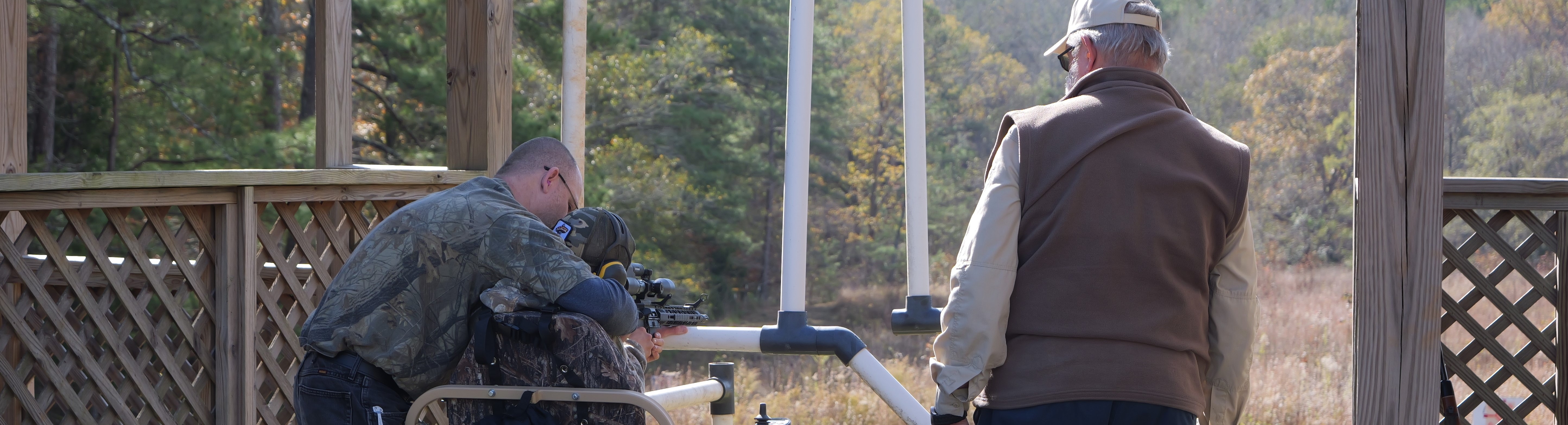 A man in a wheelcheer is assisted while shooting a gun.